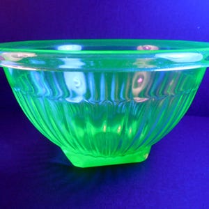 May include: A vibrant, glowing green glass bowl with a ribbed exterior. The bowl has a wide, rounded rim and a slightly squared base. The glass appears translucent, with light reflecting off the ridges.