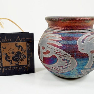 May include: A hand-thrown Raku art pottery vase with a Kokopelli design. The vase has a metallic finish with shades of red, blue, and silver. A black tag with the words "Raku Art" and "Authentic Kokopelli" hangs nearby.