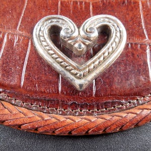 May include: A brown leather accessory with a crocodile-embossed texture and a braided leather border. Centered on the surface is a polished silver-toned metal heart ornament featuring intricate scrollwork and a twisted rope-like border design.