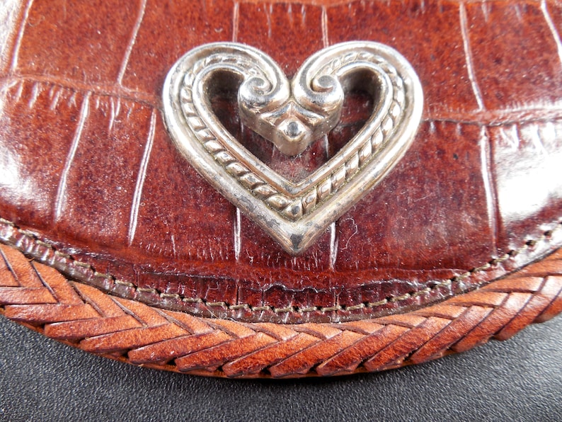 May include: A brown leather accessory with a crocodile-embossed texture and a braided leather border. Centered on the surface is a polished silver-toned metal heart ornament featuring intricate scrollwork and a twisted rope-like border design.