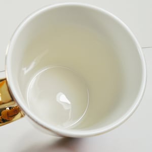 May include: A cream-colored ceramic mug with a gold-colored handle. The mug has a glossy finish and a simple, elegant design. The interior of the mug is also cream-colored.
