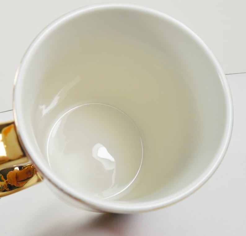 May include: A cream-colored ceramic mug with a gold-colored handle. The mug has a glossy finish and a simple, elegant design. The interior of the mug is also cream-colored.