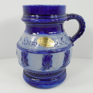 May include: A blue and white ceramic stein with a handle. The stein has a raised design of a rooster and the text "Gres D'Alsace Fait Main Ed. Schmitt 67 Betschdorf".