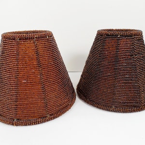 May include: Two brown beaded lamp shades with a tapered shape. The shades are made of small, dark brown beads, giving them a textured appearance. The shades are designed to fit over a light bulb and diffuse light.