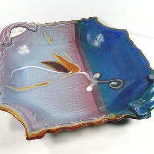 May include: A handmade ceramic serving dish with a unique, asymmetrical shape. The dish features a color gradient from purple to blue, with a brown and white floral design. It has a scalloped edge and handles.