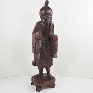 May include: A carved wooden statue of an elderly man with a long beard and traditional clothing. The statue is dark brown and stands on a carved base. The man is holding a lantern.