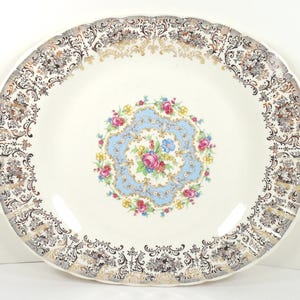 May include: A vintage white ceramic serving platter with scalloped edges and two handles. The rim features a gold and black floral pattern. The center has a light blue design with a floral bouquet.