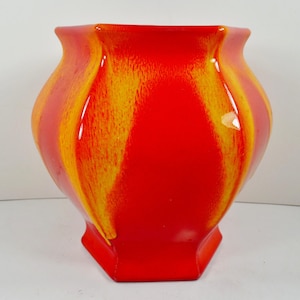 May include: A vibrant, red ceramic vase with vertical yellow and orange streaks. The vase has a unique, geometric shape with a wide opening and a hexagonal base. The glossy finish enhances the bright colors, making it a striking decorative piece.