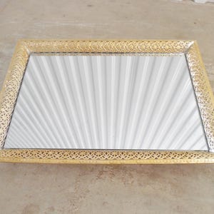 May include: Rectangular gold-toned mirror with an ornate, openwork frame. The mirror's surface reflects a fan-like pattern. The frame has a delicate, repeating geometric design. The mirror is a decorative item.