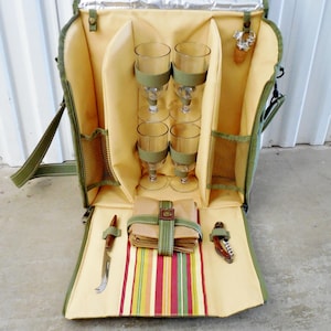 May include: Open picnic backpack with olive green trim and a tan interior. It contains four wine glasses, a corkscrew, a knife, and a folded cloth napkin set. The bottom has a striped pattern in red, yellow, and green.