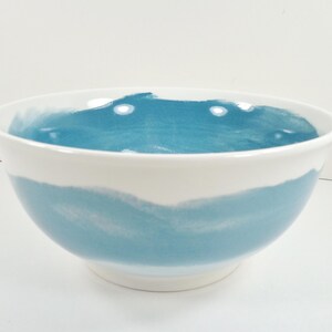 May include: A white ceramic bowl with a wide, rounded shape. The bowl features a band of light blue watercolor-style paint around the middle and inside the bowl. The bowl is suitable for serving food.