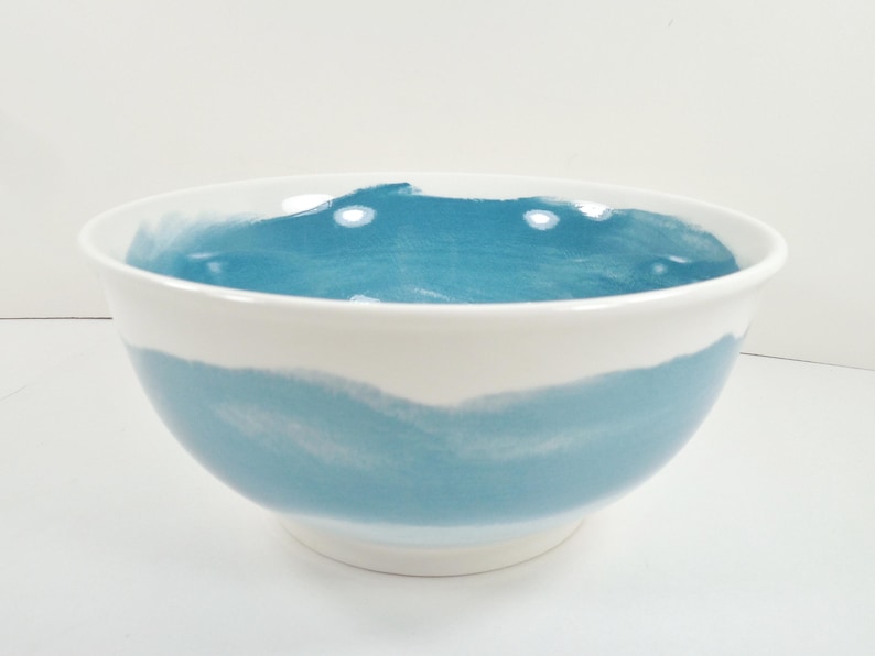 May include: A white ceramic bowl with a wide, rounded shape. The bowl features a band of light blue watercolor-style paint around the middle and inside the bowl. The bowl is suitable for serving food.
