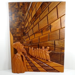 May include: Wooden relief carving depicting a religious scene. The artwork features figures in robes and head coverings, set against a backdrop of a brick wall and a mosaic-style floor. The carving is made of various shades of brown wood.