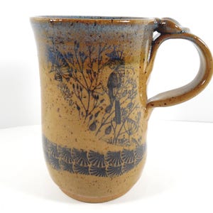 May include: A handmade ceramic mug with a brown and blue glaze. The mug features a detailed black illustration of a bird and foliage. The handle has a decorative element. The mug is approximately 5 inches tall.