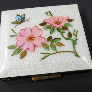 May include: Square, white compact with a floral design. Pink flowers with green leaves and a blue butterfly are painted on the textured surface. The compact has a gold-colored metal frame and clasp.