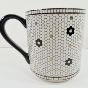May include: White ceramic mug with a black handle, featuring a honeycomb pattern with black flower and gold dot accents. The rim and base have a black and white border. A stylish and decorative coffee mug.