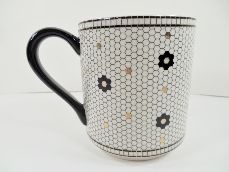 May include: White ceramic mug with a black handle, featuring a honeycomb pattern with black flower and gold dot accents. The rim and base have a black and white border. A stylish and decorative coffee mug.