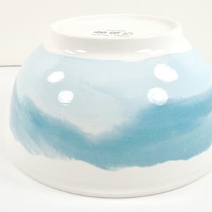 May include: A white ceramic bowl with a blue watercolour design. The bowl has a wide, rounded shape with a white rim and a blue and white painted exterior. The blue paint has a watercolour effect.