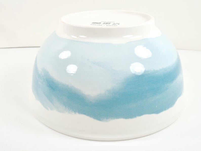 May include: A white ceramic bowl with a blue watercolour design. The bowl has a wide, rounded shape with a white rim and a blue and white painted exterior. The blue paint has a watercolour effect.