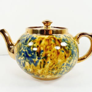 May include: A ceramic teapot with a rounded body, a gold-colored spout, handle, and lid. The teapot features a speckled pattern in shades of blue, yellow, and brown. The lid has a gold finial. A classic tea accessory.