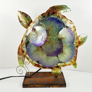 May include: A decorative metal sea turtle sculpture with a multi-colored shell. The shell features iridescent blue, green, and purple circular designs with gold accents. The sculpture is mounted on a rectangular, rusty-brown base.