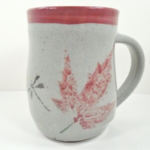 May include: A handmade ceramic mug with a gray speckled finish. The rim is painted red, and a large red leaf and a dragonfly are painted on the side. The mug has a curved handle.