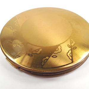 May include: A vintage, round, gold-toned compact with etched floral and leaf designs. The compact has a hinged opening and a slightly worn appearance, suggesting age and use. The design is symmetrical, with the etched details adding visual interest.