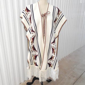 May include: An off-white poncho featuring vertical black stripes and a repeating floral pattern in shades of red, brown, and green. The garment includes a braided cord tie at the neckline and a long, white fringe along the bottom hemline.