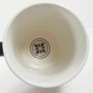 May include: White ceramic mug with a black handle. The interior features a black and white floral design at the bottom. The mug is empty and appears clean, with a glossy finish.