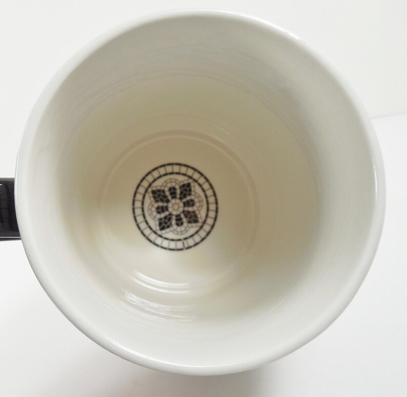 May include: White ceramic mug with a black handle. The interior features a black and white floral design at the bottom. The mug is empty and appears clean, with a glossy finish.