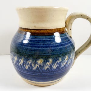May include: A handmade ceramic pitcher with a wide, rounded body and a sturdy handle. The pitcher features a cream-colored rim and handle, a dark brown band, and a blue body with a decorative white and blue zigzag pattern. The pitcher is approximately 7 inches tall.