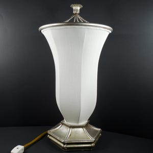 May include: A white, vase-shaped table lamp with a silver-colored base and trim. The lamp has a fabric shade and a decorative finial at the top. The lamp is plugged into an electrical outlet.