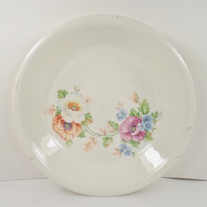 May include: A round, off-white ceramic bowl featuring a central floral decal. The design displays a cluster of flowers in shades of orange, white, pink, and blue, accented with green leaves and stems, arranged in a curved pattern across the center.