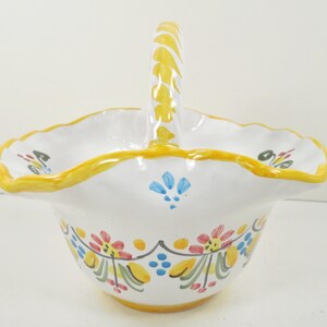 May include: A white ceramic basket with a yellow handle and rim. The basket is decorated with colorful floral and leaf patterns in red, blue, and yellow. The handle has a yellow, textured design.