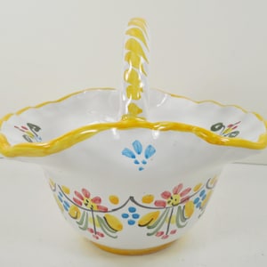 May include: White ceramic basket with a yellow handle and rim. The basket is decorated with colorful floral patterns in red, yellow, blue, and green. The handle has a twisted design.