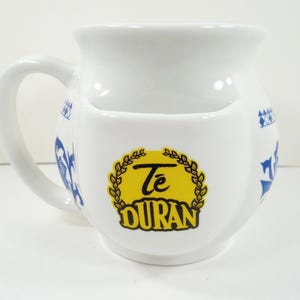 May include: White ceramic mug with a built-in pocket, featuring blue decorative patterns and the text "Té Duran" in a yellow emblem. The mug has a curved handle and a wide opening.