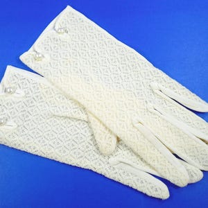 May include: A pair of off-white, open-weave gloves with a diamond pattern. The gloves have a scalloped edge and are adorned with three faux pearls on each cuff. The background is a solid blue.