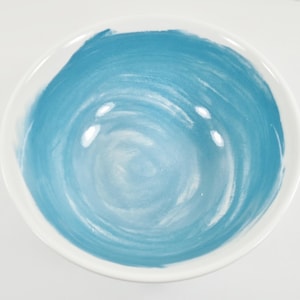 May include: A white ceramic bowl with a hand-painted blue swirl design in the centre. The bowl has a smooth, glossy finish and is suitable for serving food or as a decorative piece.