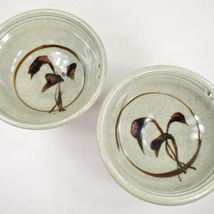 May include: Two ceramic bowls with a speckled, light green glaze. Each bowl features a brown, stylized floral design within a circular border. The bowls are suitable for serving soups, salads, or other dishes.