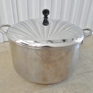 May include: A large stainless steel stockpot with a polished finish, featuring two side handles and a matching lid. The lid has a sunburst pattern radiating from the center and is topped with a small, round, black knob handle for easy lifting.