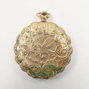 May include: Ornate gold-toned pocket watch with a detailed floral and bird design. The watch has a scalloped edge and a textured surface. The bird is in flight, and the watch has a loop at the top for a chain.