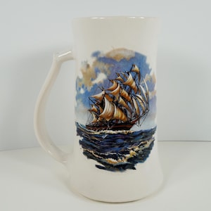 May include: White ceramic mug with a blue and white illustration of a sailing ship with brown sails. The ship is sailing on a blue ocean with white waves.