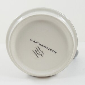 May include: Off-white ceramic pitcher with a rounded base and a handle. The bottom of the pitcher features the text "by ANTHROPOLOGIE", "Made in China", "Fabriqu&eacute; en Chine", and "Hand wash recommended".