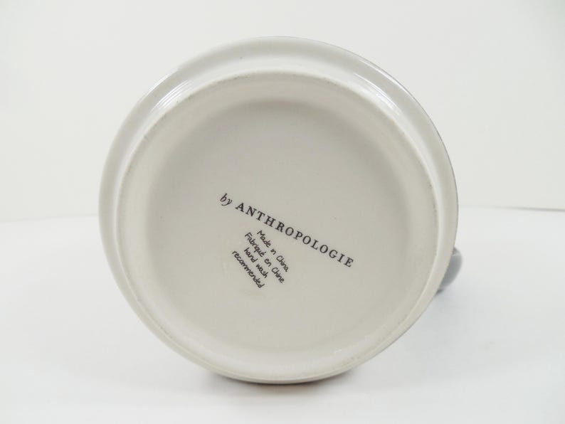 May include: Off-white ceramic pitcher with a rounded base and a handle. The bottom of the pitcher features the text "by ANTHROPOLOGIE", "Made in China", "Fabriqu&eacute; en Chine", and "Hand wash recommended".