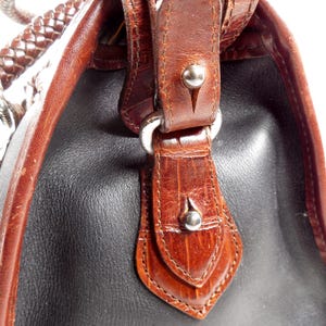 May include: A handbag featuring a combination of smooth black leather and brown textured leather panels. The bag includes a braided brown leather handle, silver-toned metal hardware, and decorative leather tabs secured with small silver studs.