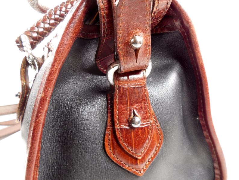 May include: A handbag featuring a combination of smooth black leather and brown textured leather panels. The bag includes a braided brown leather handle, silver-toned metal hardware, and decorative leather tabs secured with small silver studs.