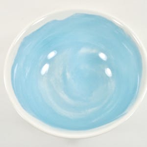May include: A round ceramic bowl with a white rim and a light blue interior. The blue paint has a swirled pattern. The bowl is empty and sits on a white surface.