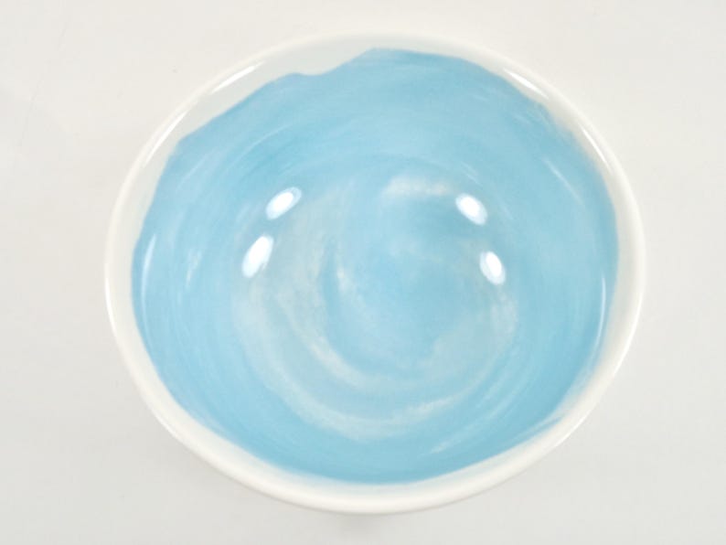 May include: A round ceramic bowl with a white rim and a light blue interior. The blue paint has a swirled pattern. The bowl is empty and sits on a white surface.