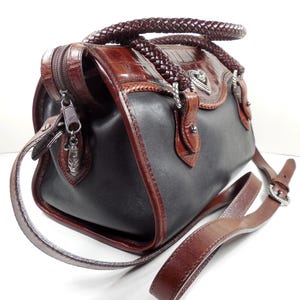 May include: A two-tone handbag featuring smooth black leather panels contrasted with brown crocodile-embossed leather trim. It includes braided leather handles, a silver-tone heart-shaped emblem, a top zipper closure, and an adjustable brown leather shoulder strap.