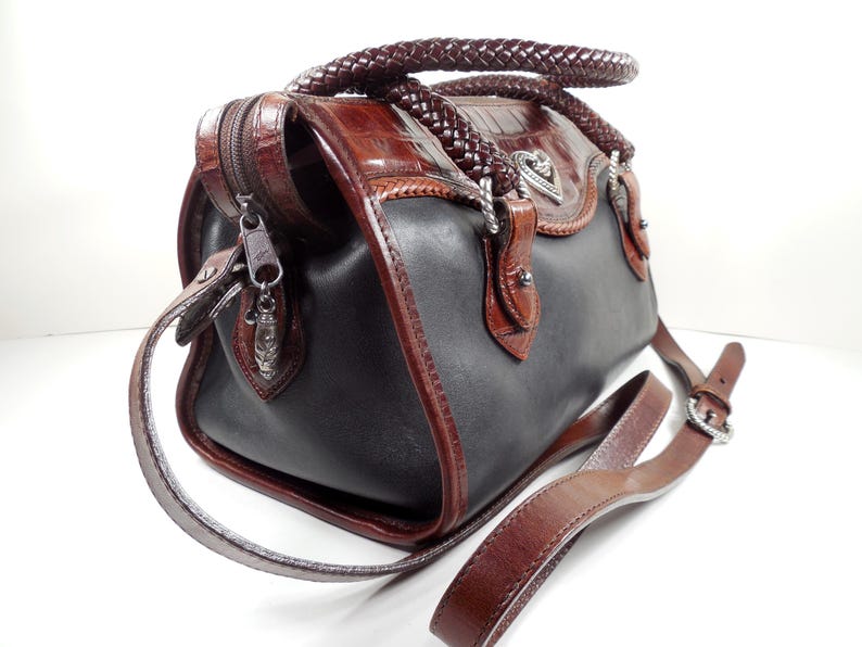 May include: A two-tone handbag featuring smooth black leather panels contrasted with brown crocodile-embossed leather trim. It includes braided leather handles, a silver-tone heart-shaped emblem, a top zipper closure, and an adjustable brown leather shoulder strap.